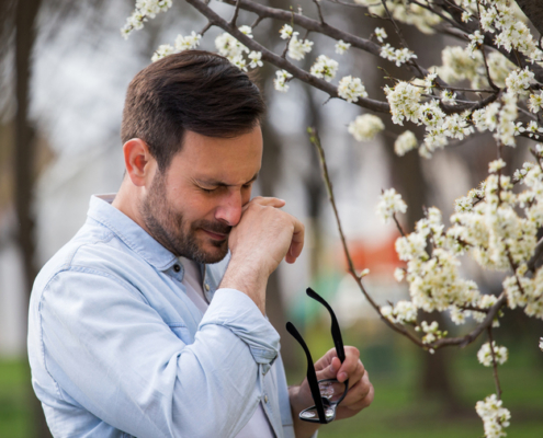 An image of a man with seasonal allergies wiping his nose beside a flowing plant.